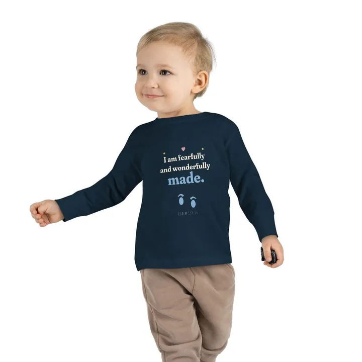 Smiling toddler wearing a navy long sleeve T-shirt with the I am fearfully and wonderfully made design with a pair of baby footprints, Psalm 139:14